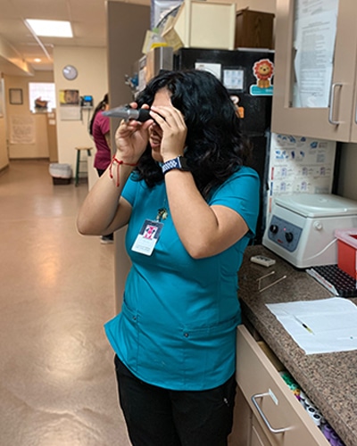 A woman in teal scrubs stands in a medical office, looking through an optical instrument. The veterinarian has dark hair, wears an ID badge, and is near a counter with lab equipment and papers.